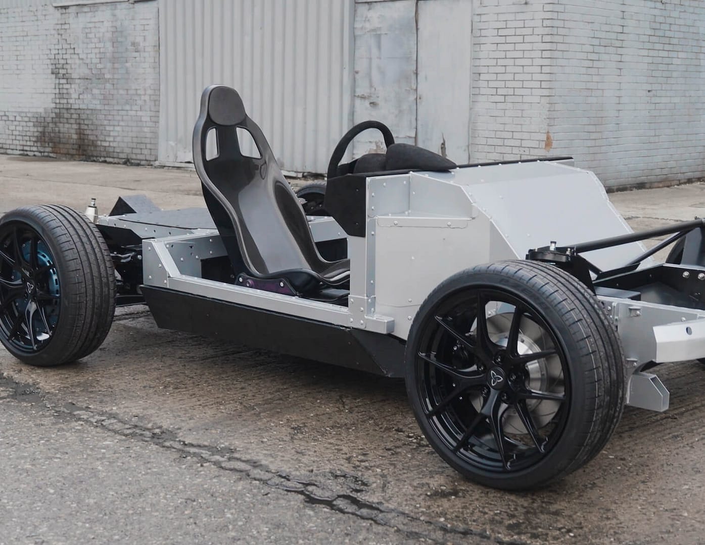 A minimalist car chassis with four wheels, a steering wheel, and two racing-style seats, shown outdoors on a concrete surface near a gray industrial building. No body panels or exterior features are present.